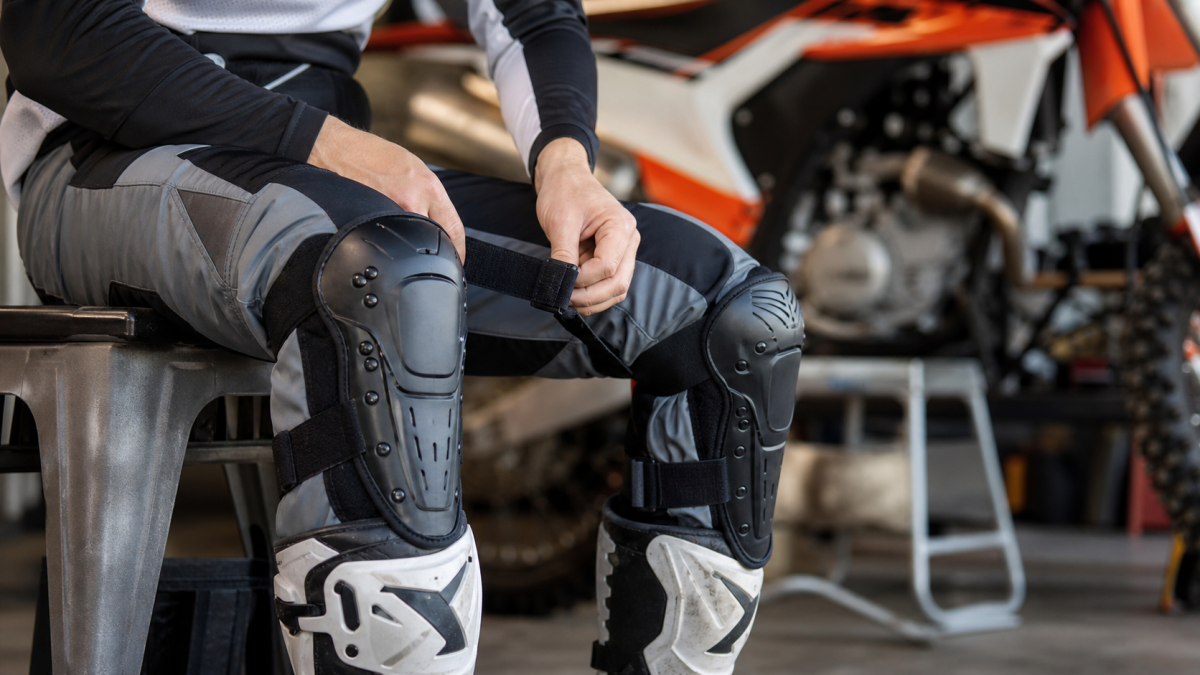 Motocross knee guards being fitted over riding pants beside a dirt bike